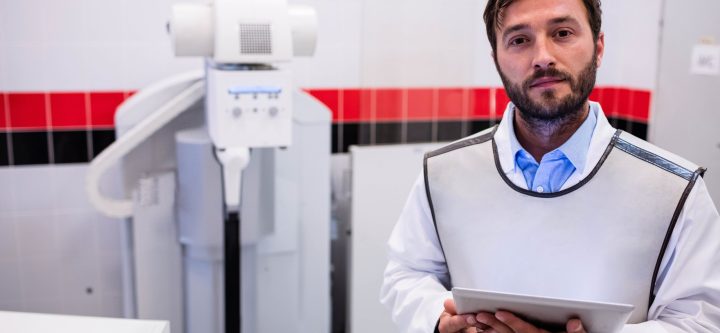 Male radiology technician wearing lead apron at x-ray machine holding tablet in clinic, copy space Male radiology technician wearing lead apron at x-ray machine holding tablet in clinic, copy space. Medical, healthcare, diagnostic, technology, clinical, professional, sterile